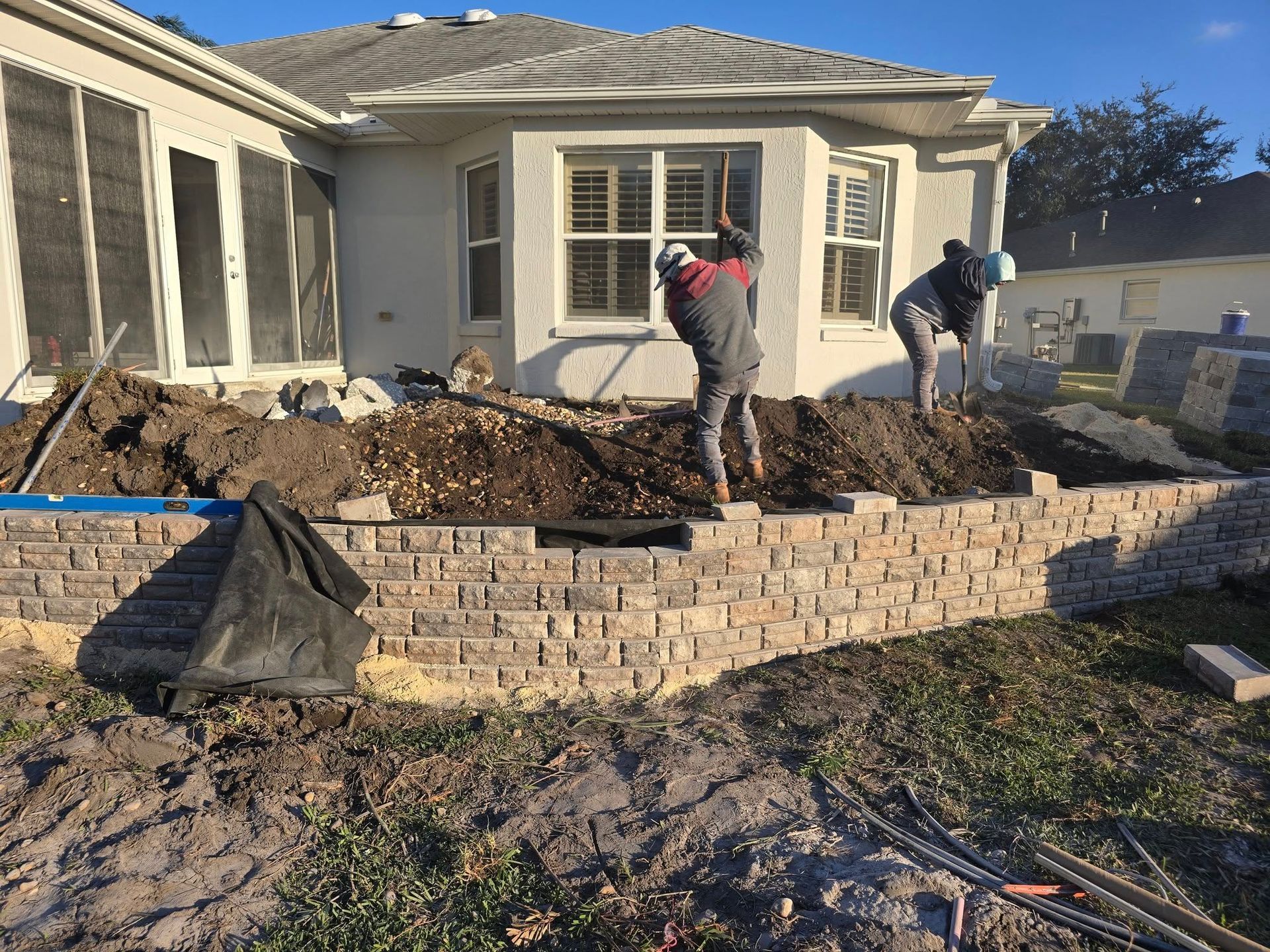 Two workers build a brick retaining wall in a residential backyard with a mound of dirt behind them.