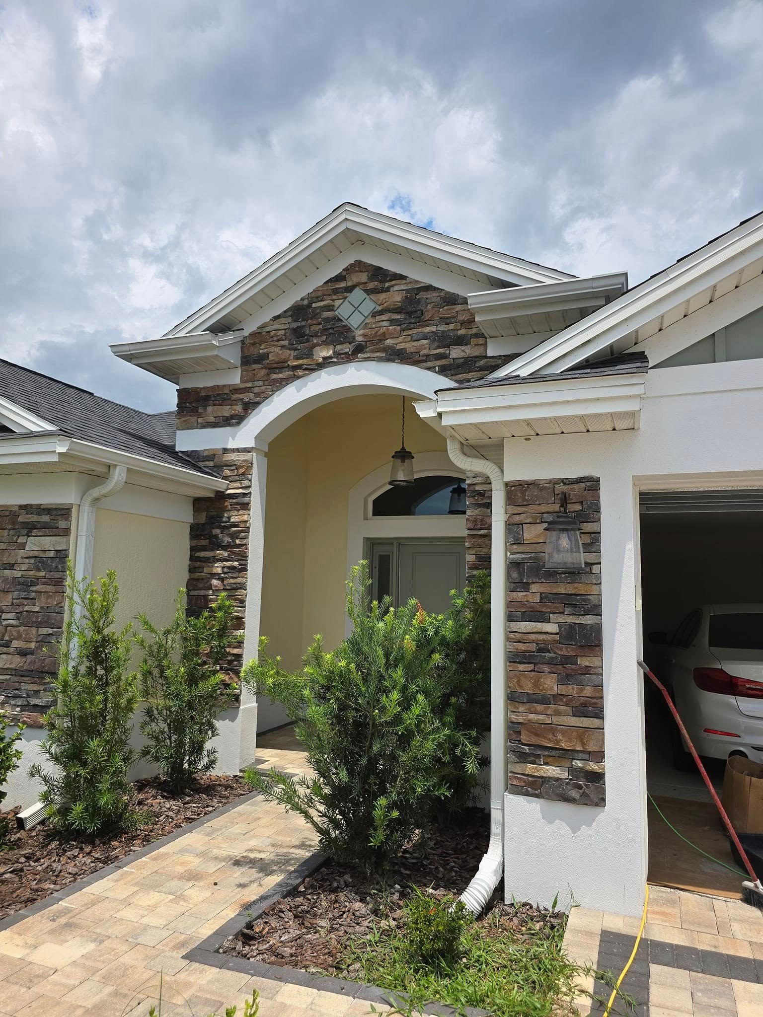 A front entrance to a single-story house with tan stucco walls, stone facade accents, a paved walkway, and landscaping.