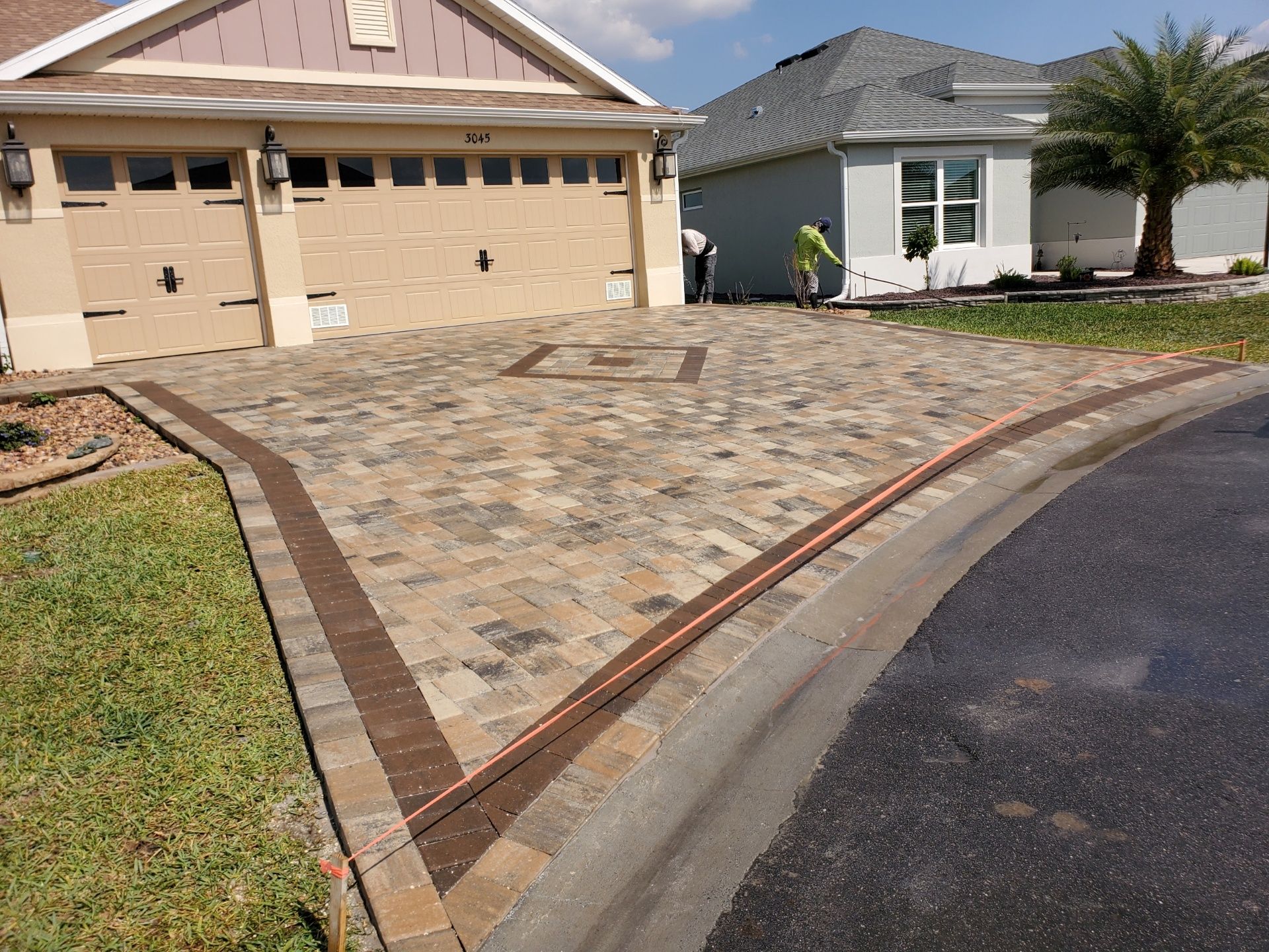 A residential driveway with patterned, multi-colored brick pavers, bordered by a darker row of bricks and a curb.
