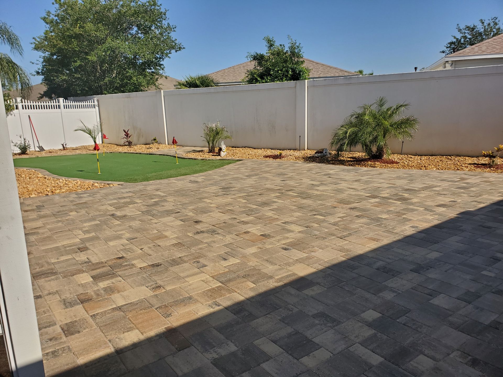 A backyard patio featuring patterned light brown pavers, a small artificial turf putting green, and a white perimeter wall.