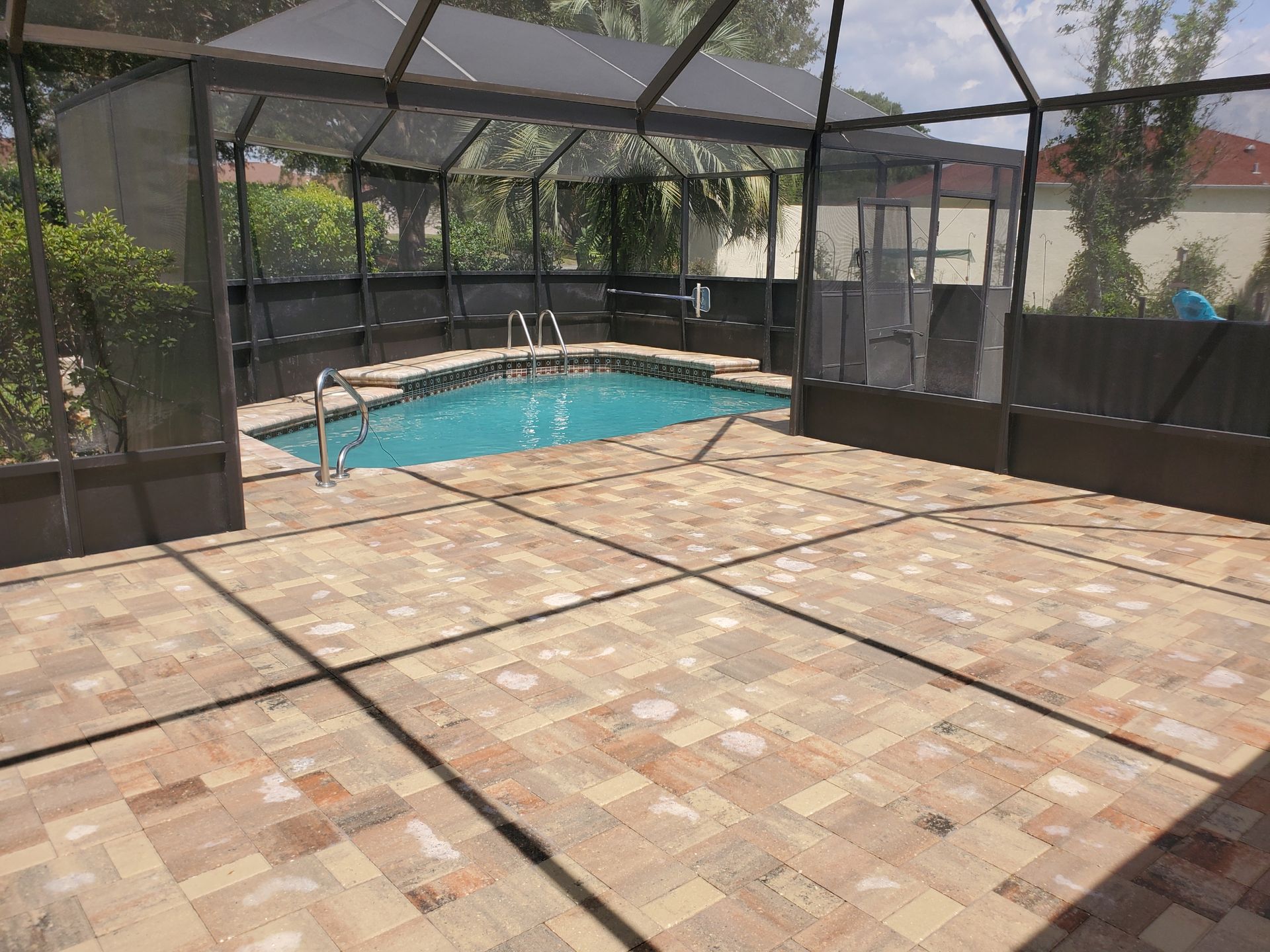 A swimming pool enclosed by a screen cage sits behind a large, tan-colored paver patio on a sunny day.