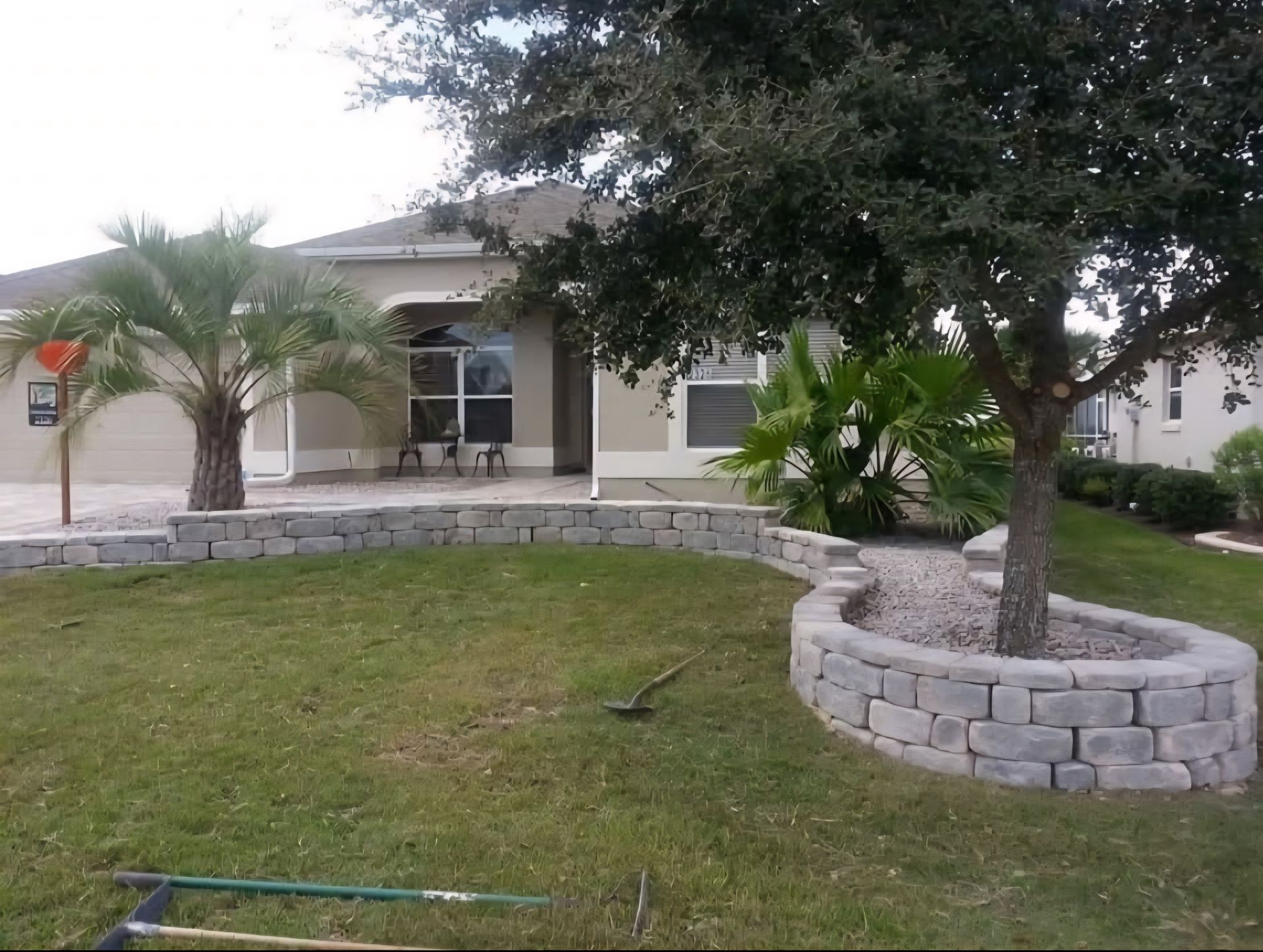 A light-colored house with a landscaped front yard featuring a low, curved stone retaining wall and a tree.