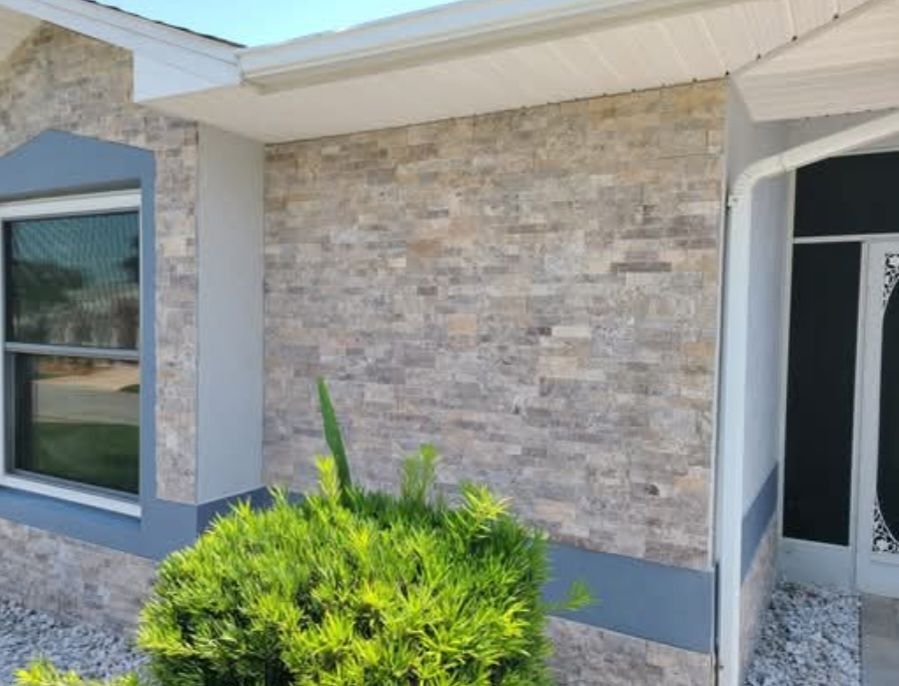 A stone-veneered house exterior with a window, dark grey trim, and a bright green bush in the foreground.