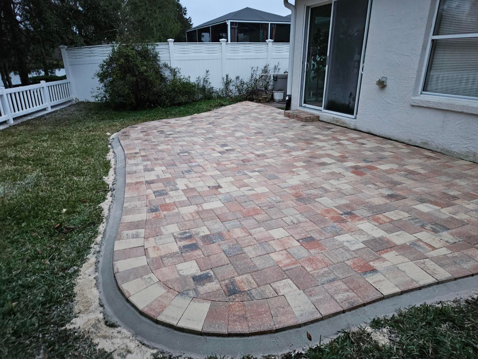 A backyard patio made of multi-colored, rectangular pavers with a smooth concrete border, next to a house.