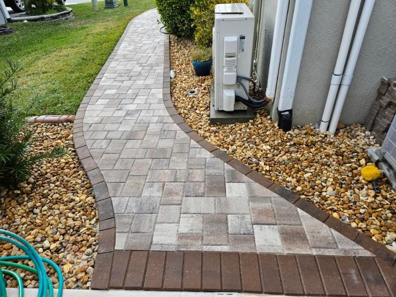 A winding paver walkway with a dark border runs between a grassy lawn and a house exterior with gravel landscaping.