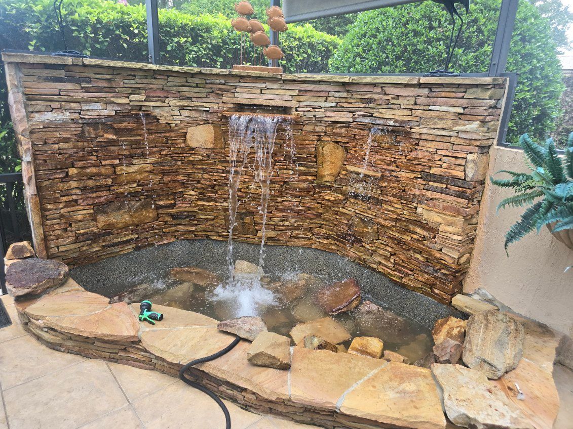A curved, stacked-stone water feature flows into a small, rock-lined basin on a patio.