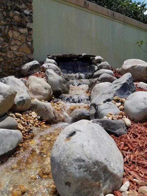 A stone water feature trickles down tiered rocks into a small stream, set against a wall and surrounded by mulch.