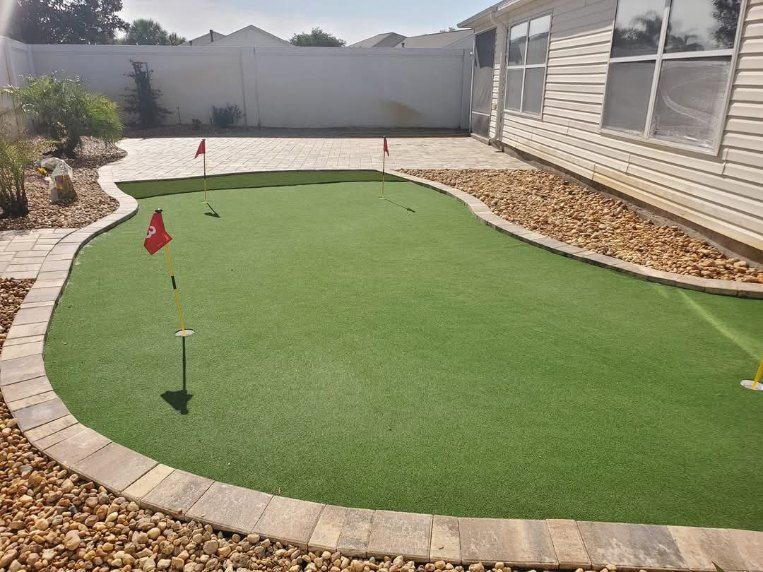 Backyard putting green with artificial grass, paver border, and three golf flags in a residential setting.