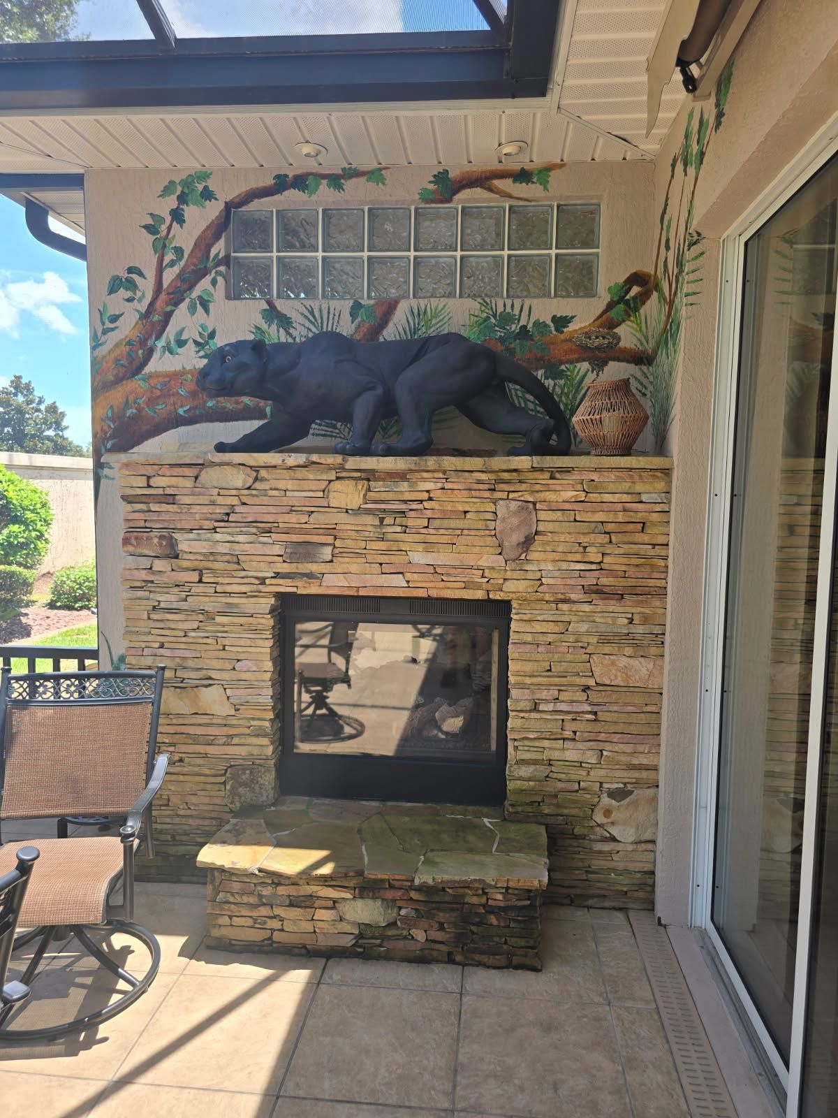 A stone fireplace on a patio featuring a black panther statue on the mantle in front of a painted jungle mural.