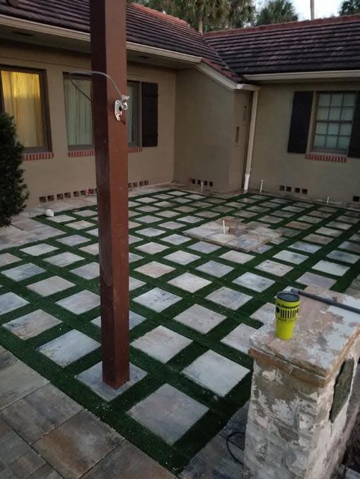 A paved patio with a grid of light gray square stones separated by artificial green turf next to a tan house.