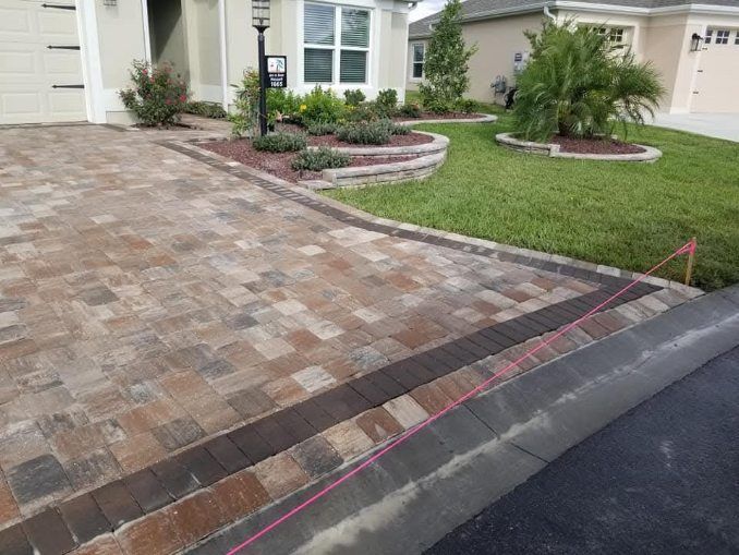 A paver driveway with a dark border meets the asphalt street in front of a house with landscaped garden beds.