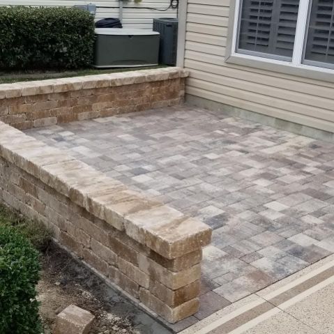 A tan, paver stone patio with a built-in seat wall against the side of a house with tan siding and a window.