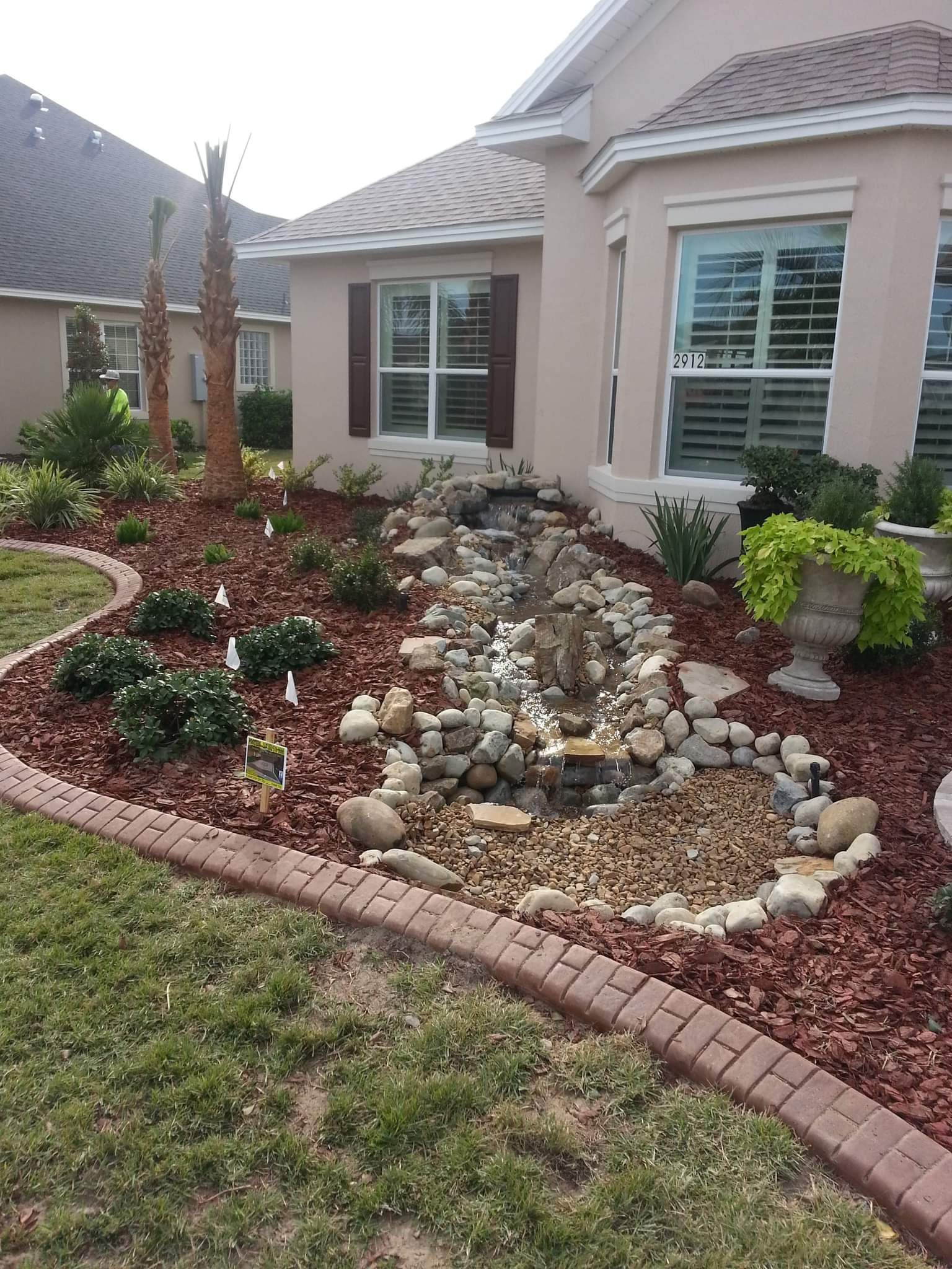 A dry creek bed of rocks flows through a mulched landscape bed in front of a tan house with brick-edged garden borders.