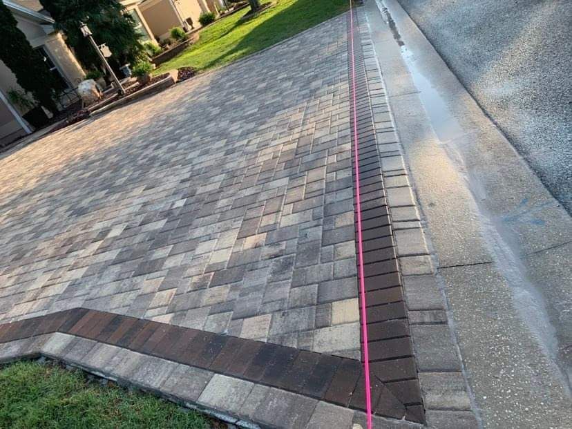 A driveway of patterned grey stone pavers with a dark brown border, bordered by a pink measuring tape and a concrete curb.