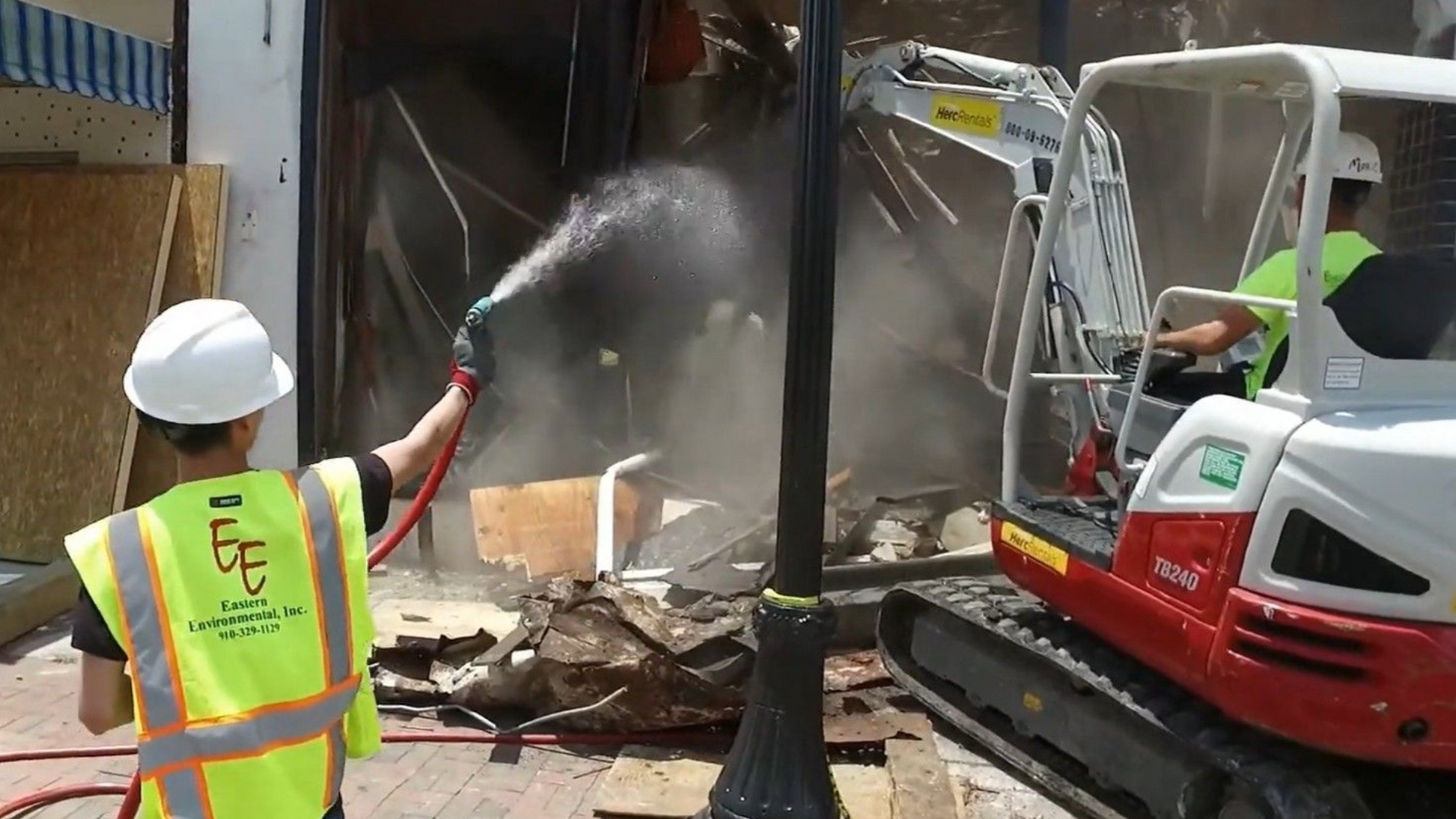 Construction workers demolishing a building storefront; one sprays water while another operates a small excavator in North Carolina.