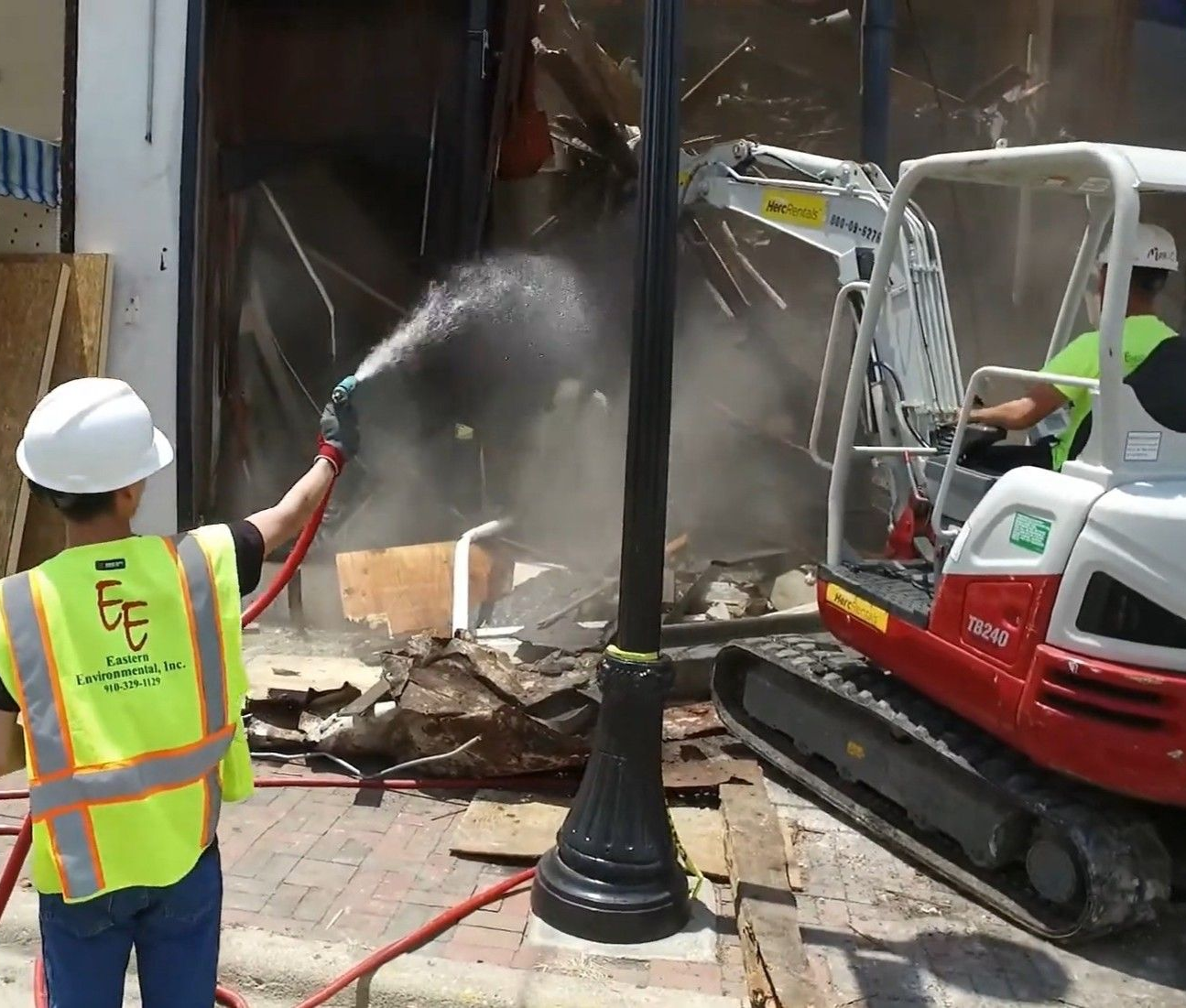 Worker spraying water at construction debris beside an excavator and streetlamp in a damaged storefront area in Nashville, TN