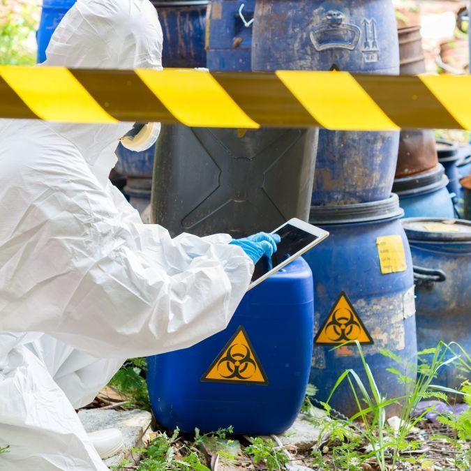 Worker in white hazmat suit checks a tablet beside biohazard-marked chemical drums and caution tape in Nashville, TN