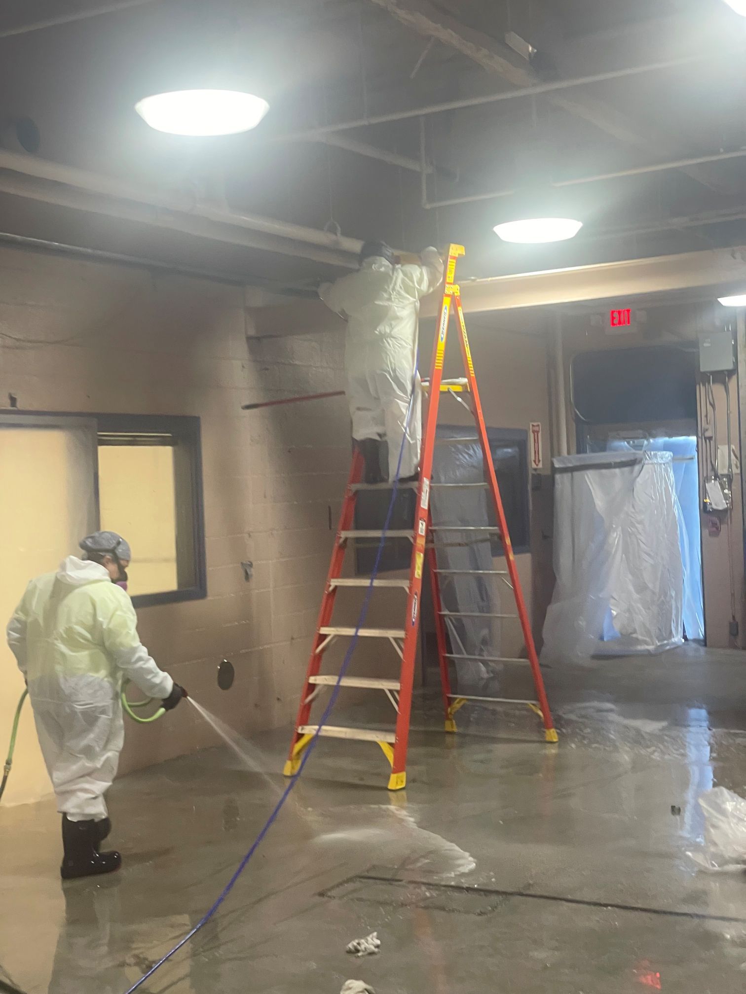 Two people in protective suits cleaning an interior space with a ladder and hose in North Carolina.