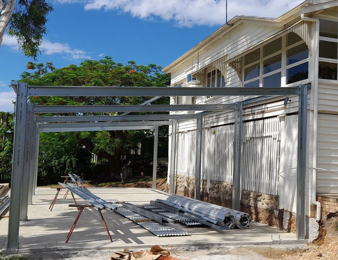 A Steel Frame Structure Being Constructed Beside a White House — BF Sheds & Garages In Rockhampton, QLD