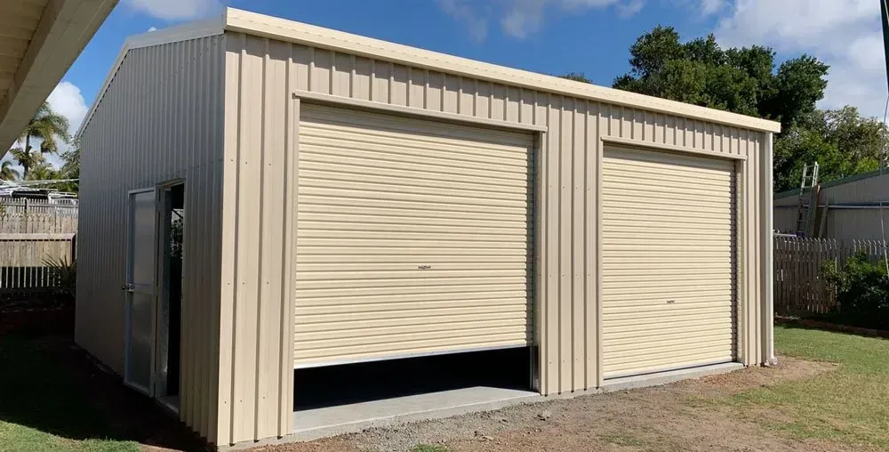 A Light Beige, Two-Car Metal Garage with Two Roll-Up Doors and A Side Entry Door, Situated in A Backyard — BF Sheds & Garages In Rockhampton, QLD