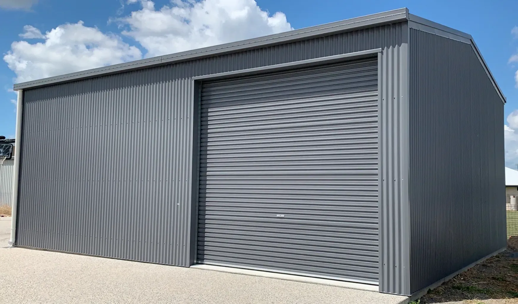 A Charcoal-Colored Metal Shed with A Roller Door, Set on A Gravel Surface Under a Partly Cloudy Blue Sky — BF Sheds & Garages In Rockhampton, QLD