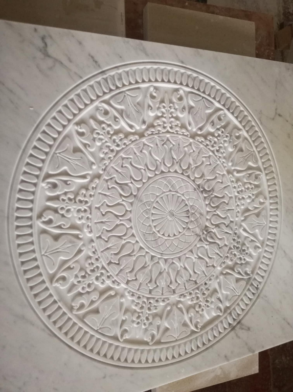 Medaglione ornamentale da parete circolare bianco con intricati rilievi floreali e geometrici su sfondo di pietra.