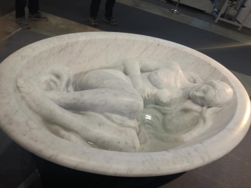 Scultura ovale bianca raffigurante due figure umane sdraiate in una vasca poco profonda, esposta in un museo.