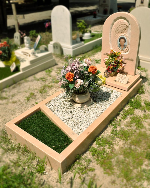 Piccolo monumento funebre color pesca con fiori e ghiaia in un cimitero, sullo sfondo di un prato verde.