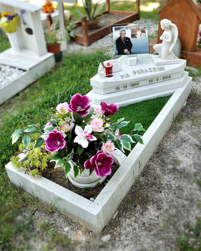 Tomba bianca con una foto incorniciata, una statua e fiori rosa in un appezzamento erboso di cimitero.