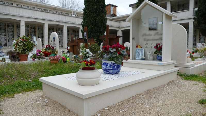 Tomba bianca con fiori e vaso blu nel giardino del cimitero sotto un cielo nuvoloso.