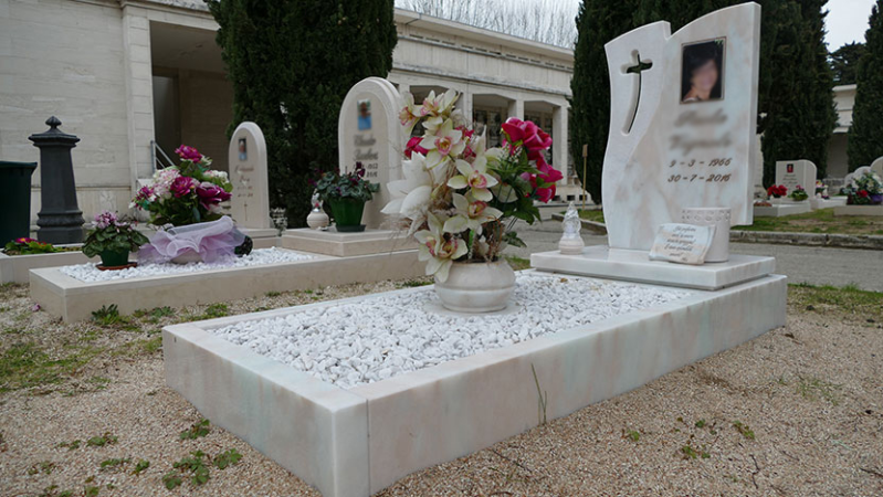 Lapide in marmo bianco con fiori in un cimitero, accanto a diverse lapidi verticali.