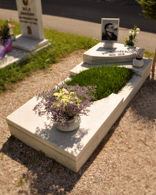 Tomba con lapidi in pietra bianca, un ritratto in bianco e nero e fiori in vaso in un cimitero soleggiato.