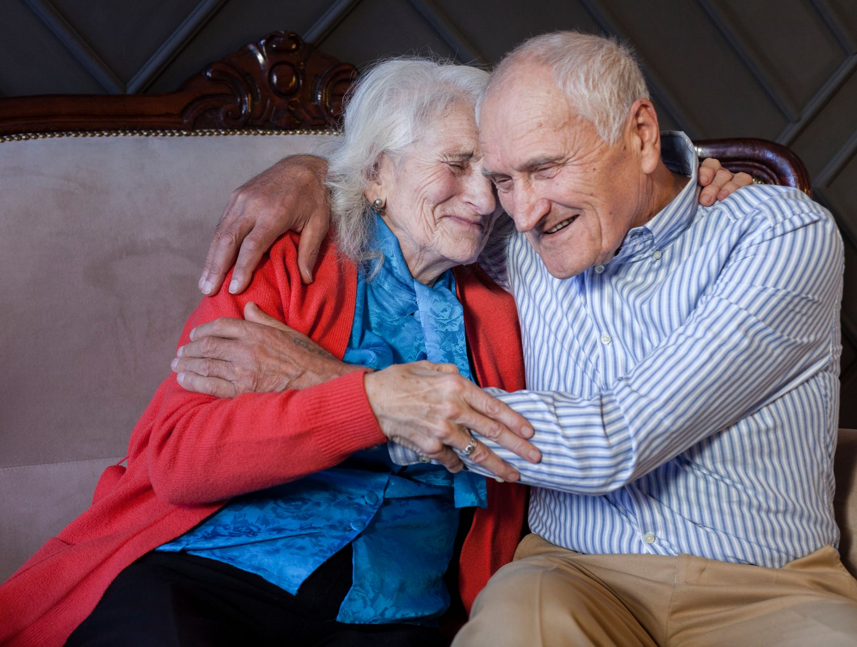 Elderly couple embraces on a sofa, smiling, and looking at each other affectionately.