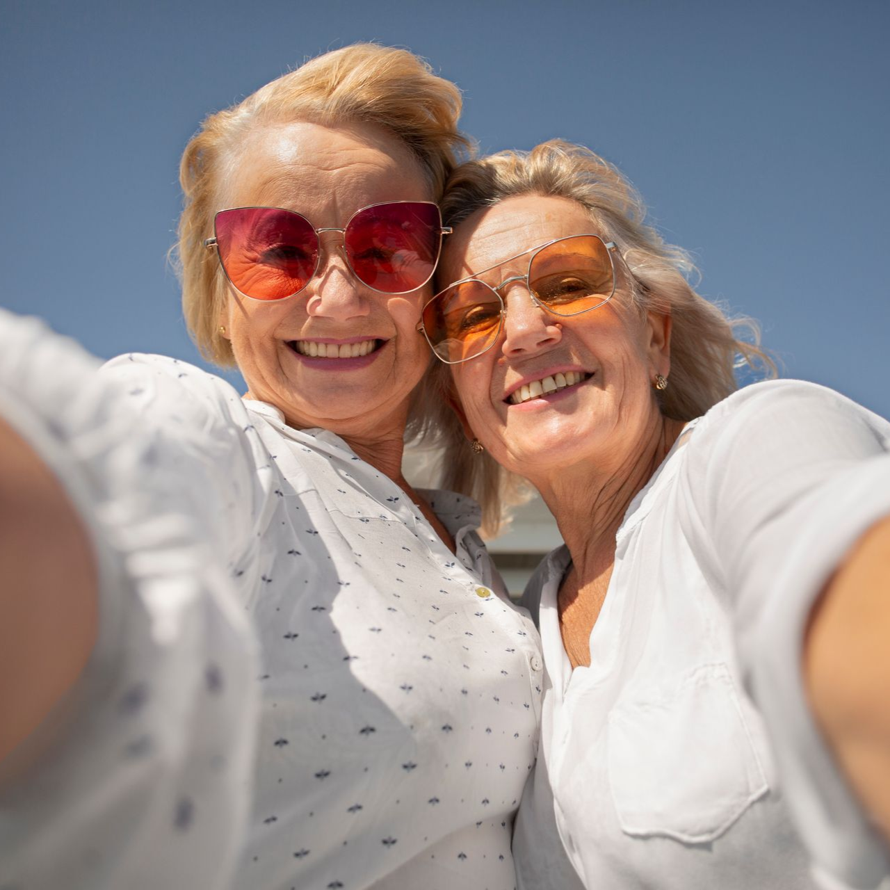 Two women smiling, taking a selfie outdoors, wearing sunglasses. Blue sky in the background.