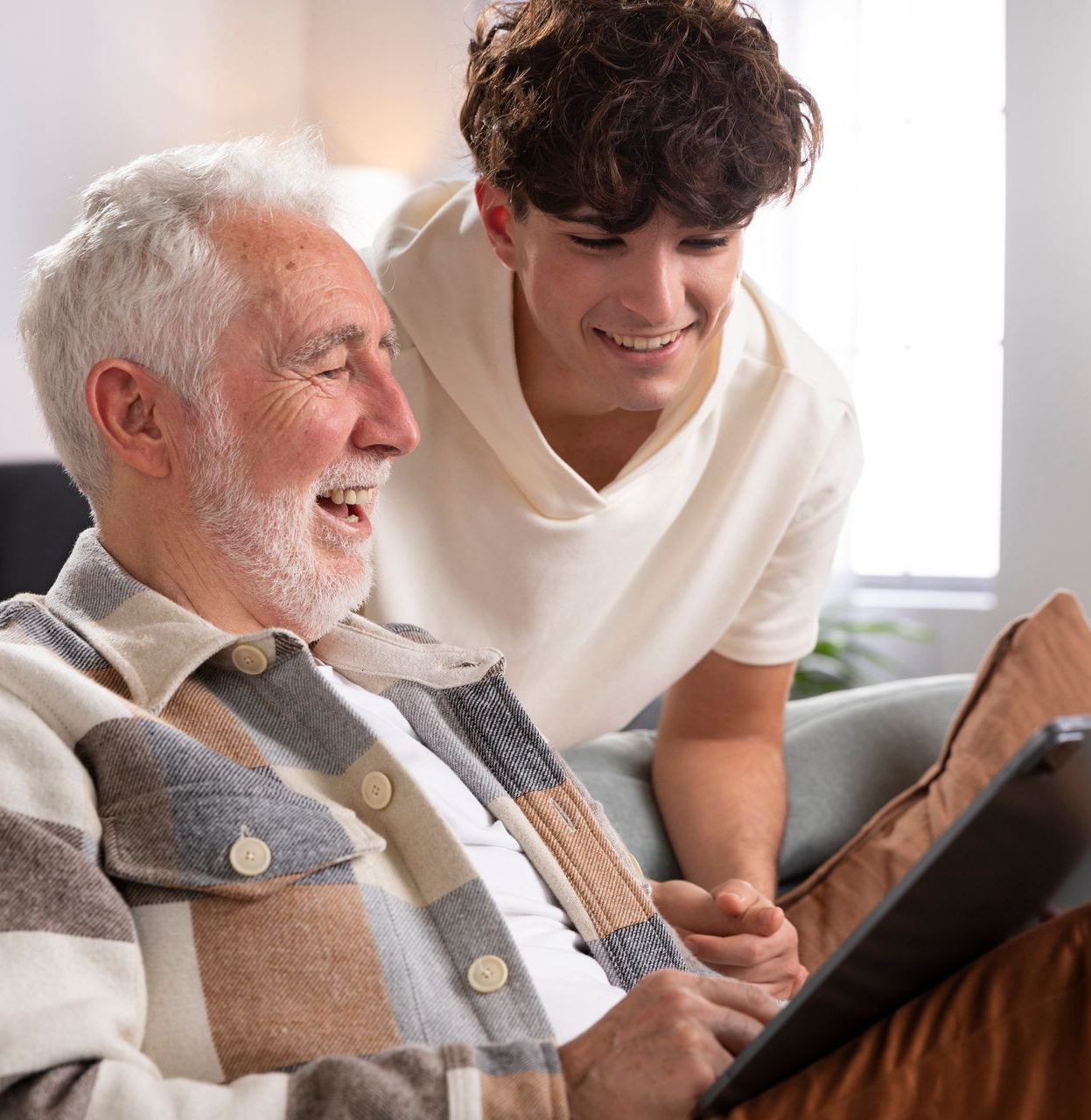 Older man and young man smiling at a tablet together, indoors.