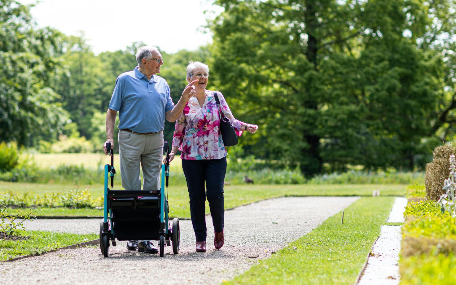 Older person with walker and another person walking on a path in a park.