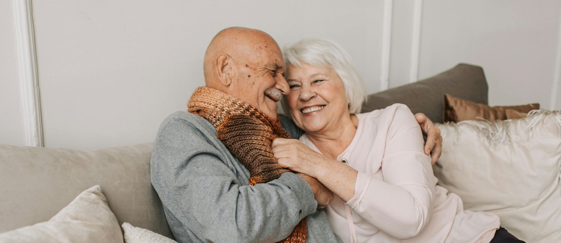 Elderly couple embracing and smiling on a sofa. Man wears a scarf. Soft, neutral background.