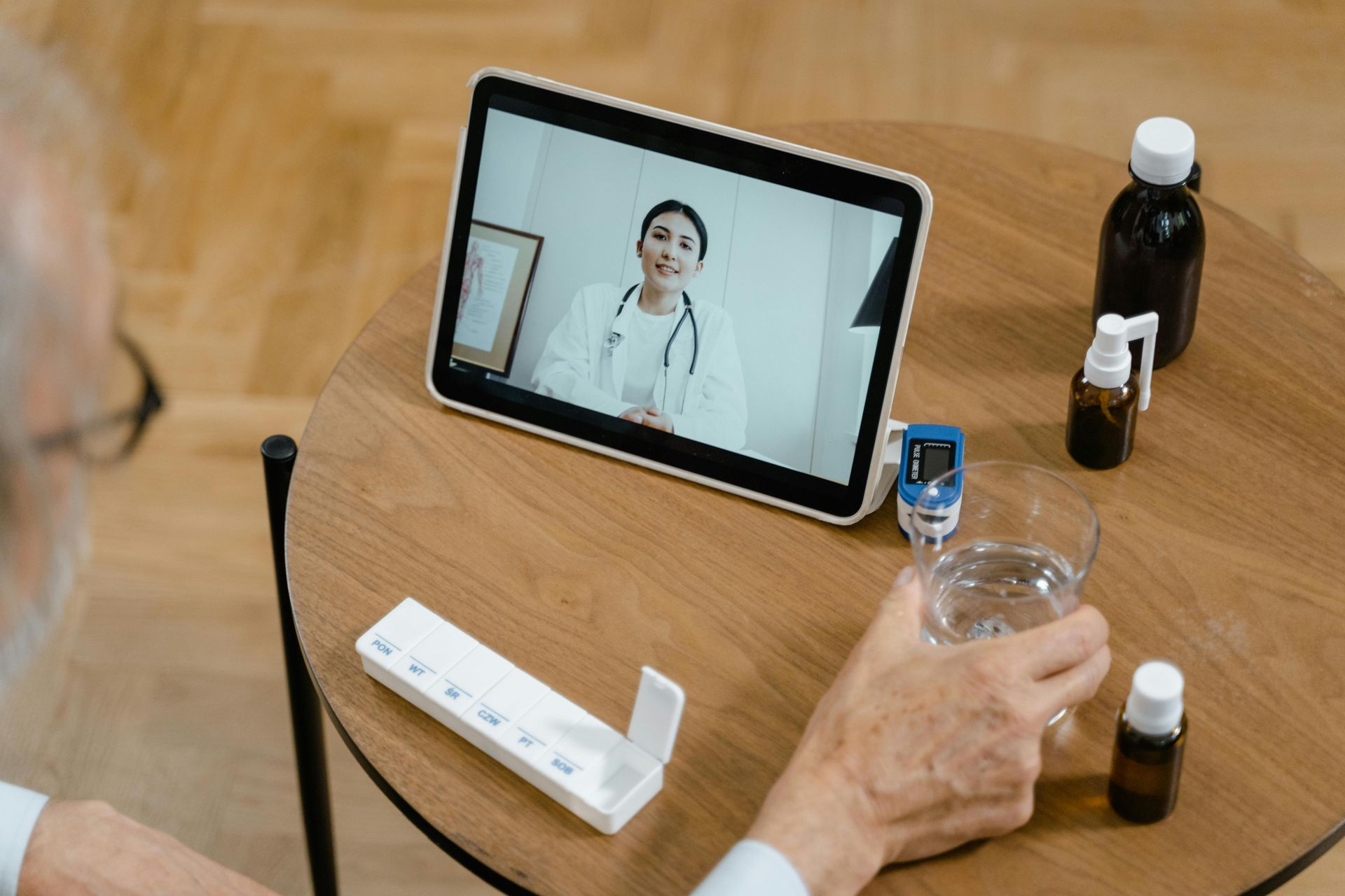 An elderly person on a telehealth call, holding a glass of water, with medication and a tablet on the table.