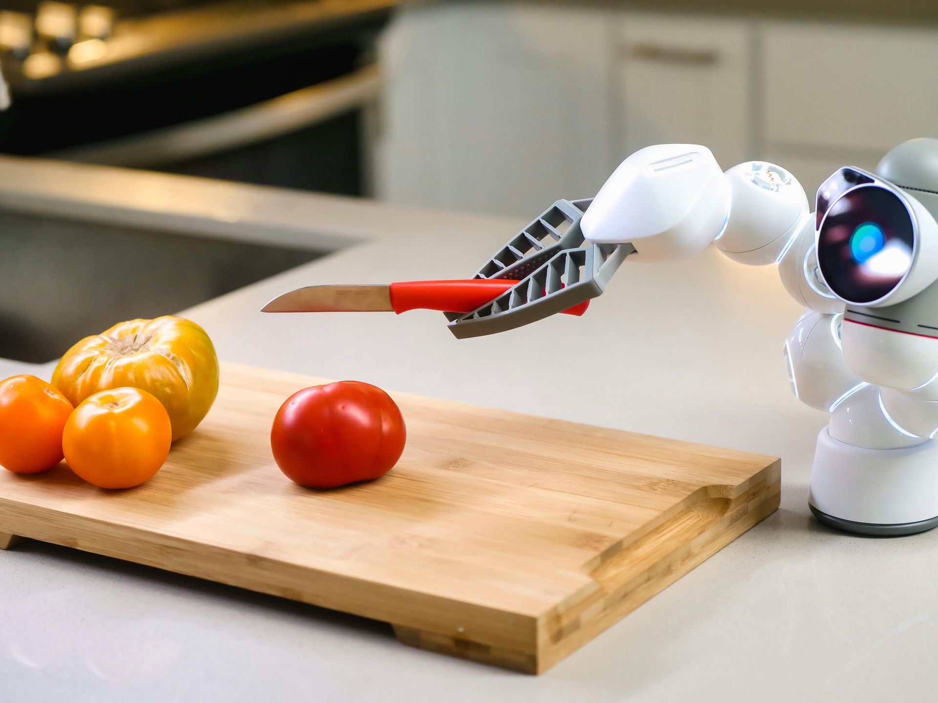 Robot arm with a knife cutting a tomato on a wooden cutting board next to other produce in a kitchen.