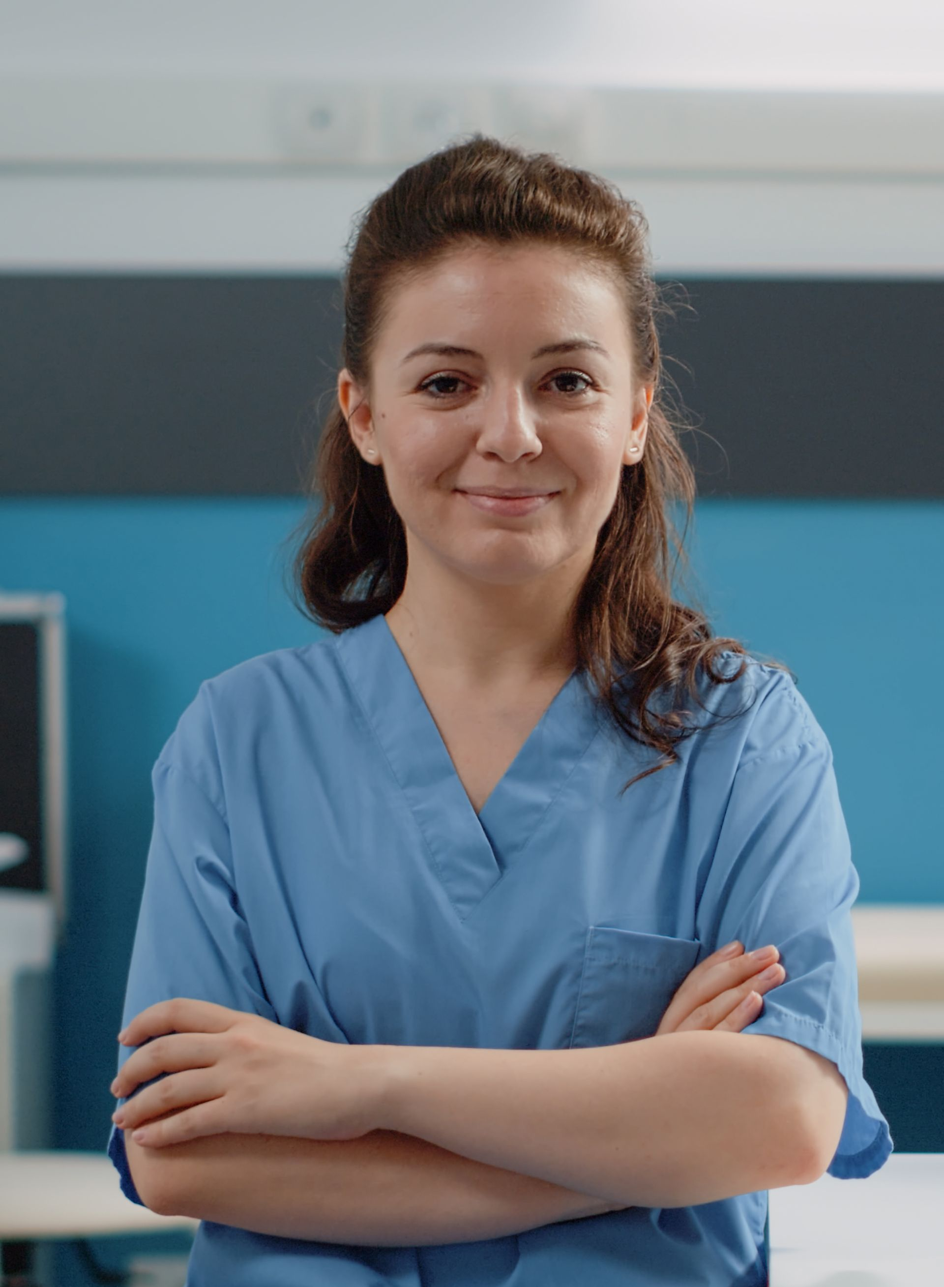 Nurse in blue scrubs, arms crossed, smiling at the camera in a medical setting with blue walls.