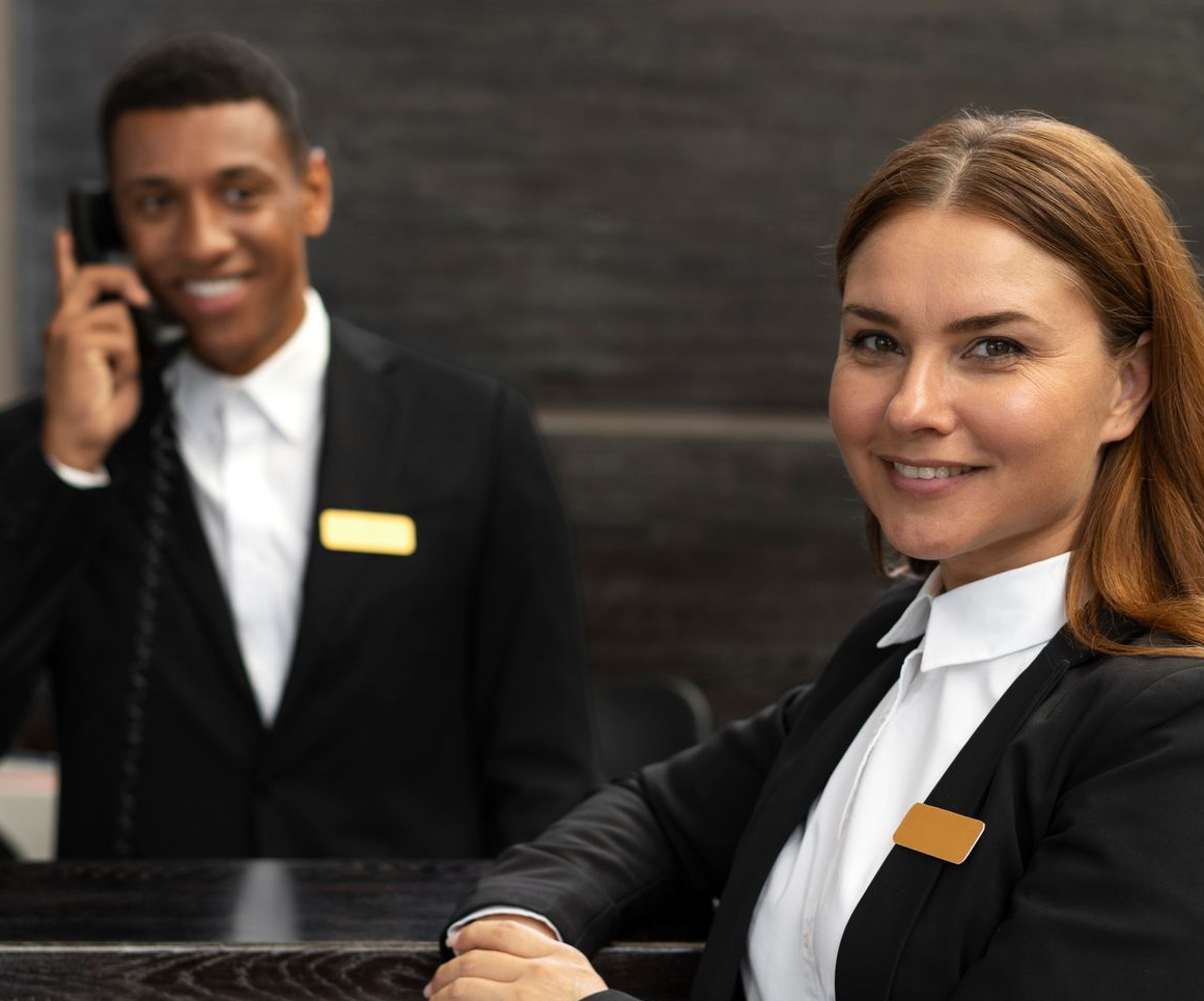 Hotel staff smiling at the camera, man on phone at the desk. Both wearing black jackets, name tags.