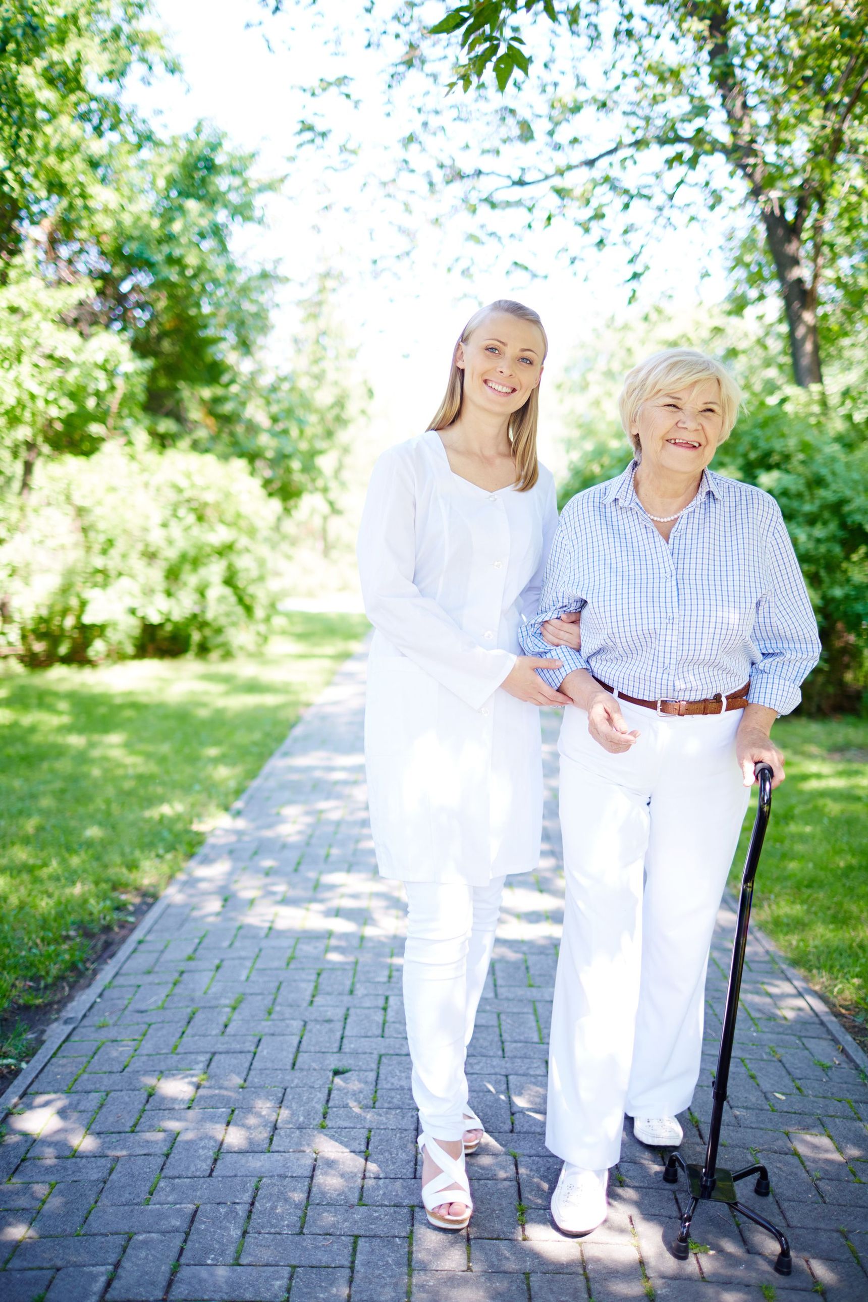 Woman assisting an older person with a cane walking on a path in a park, both smiling.