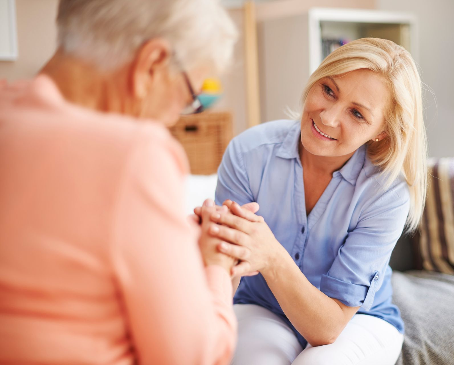 Woman holds the hands of an older person, offering comfort and support indoors; both are smiling.