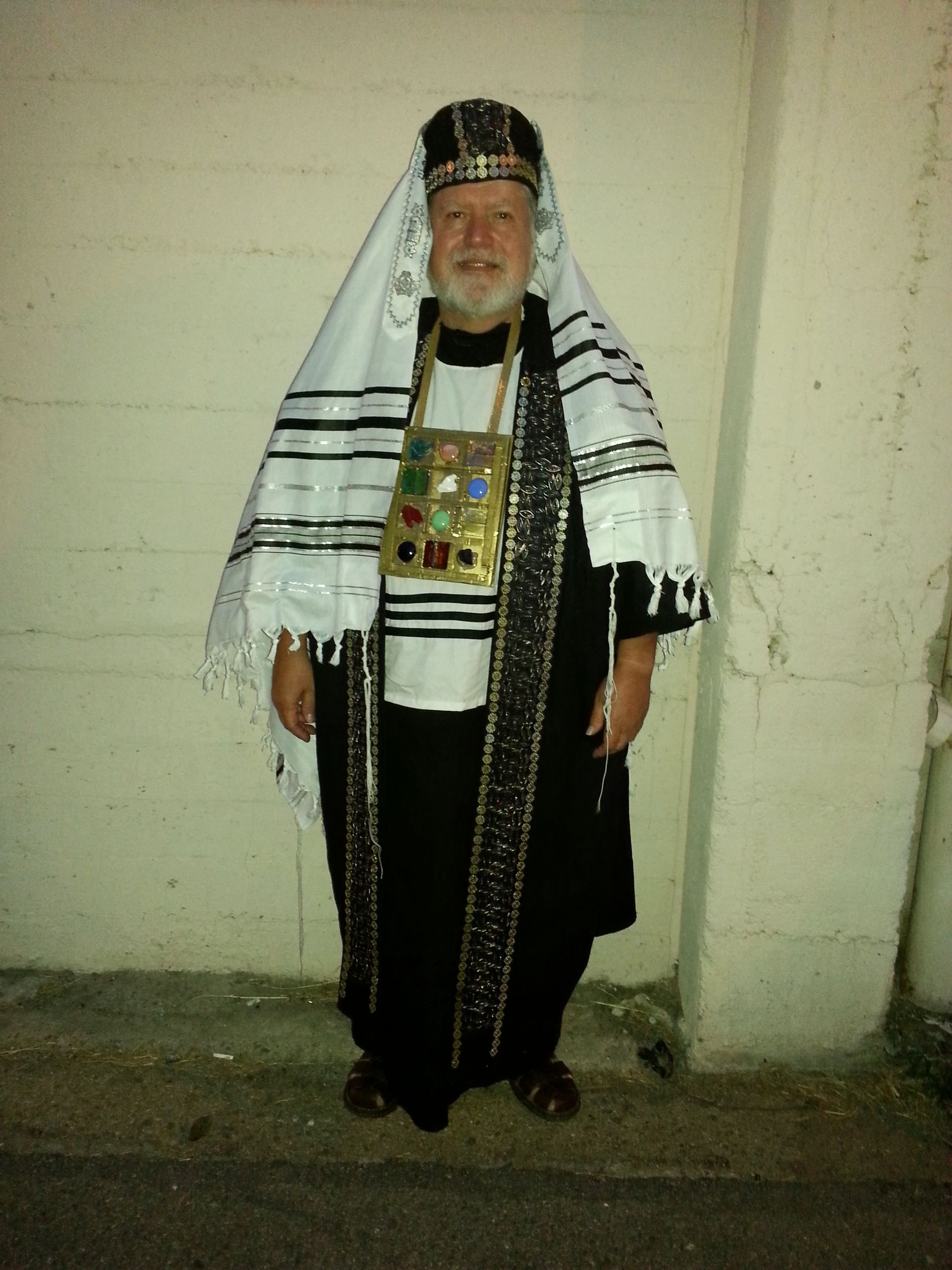 Person in traditional ceremonial clothing and head covering standing against a plain wall.