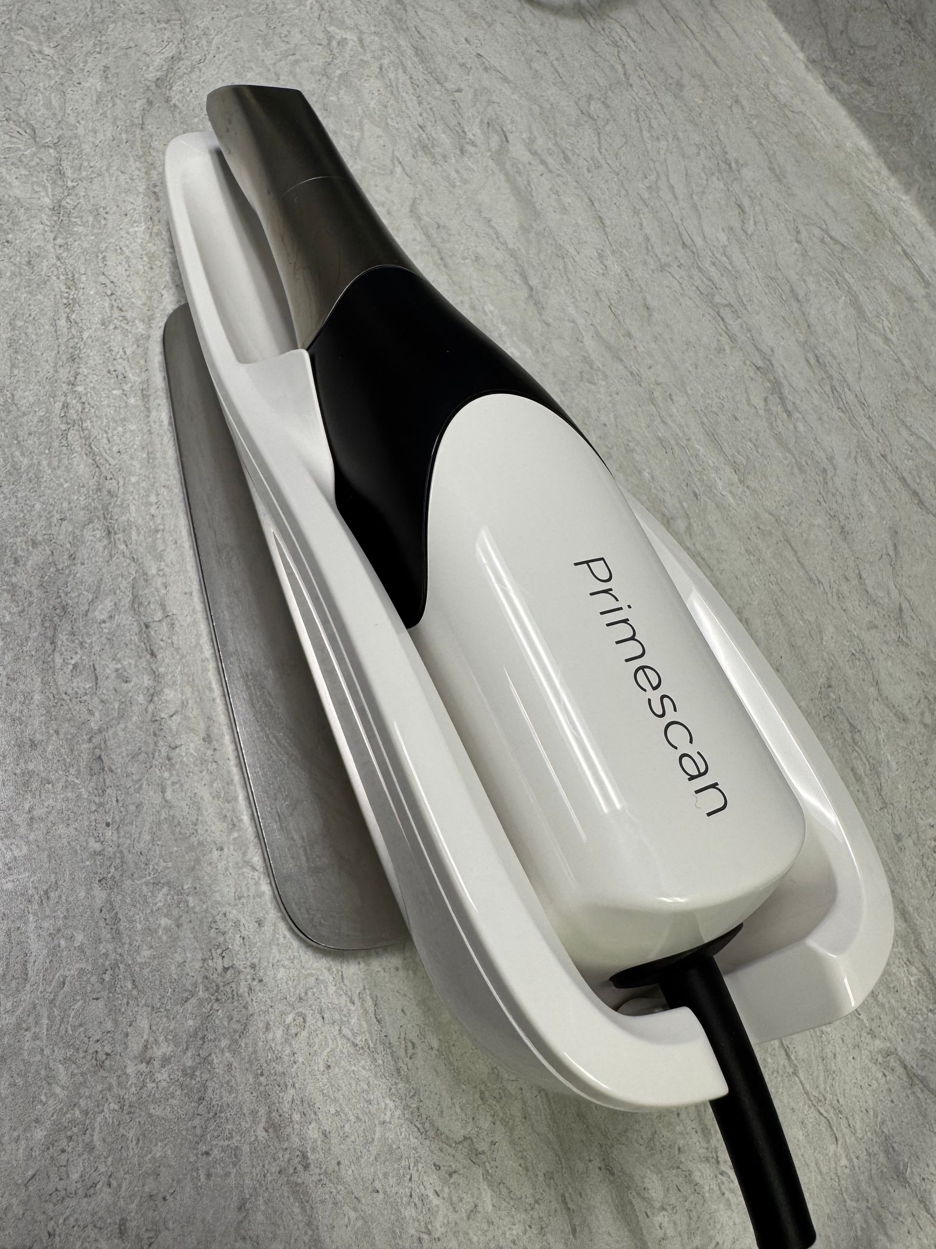 A white and black Primescan dental scanner in a white holder, resting on a gray surface.