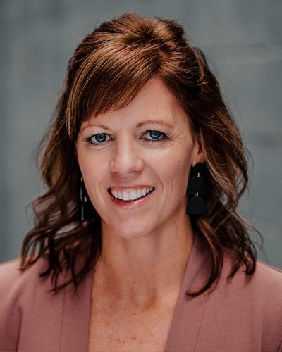 Woman with brown hair, smiling, wearing a pink blazer, black earrings.