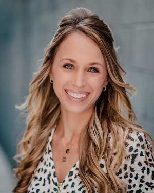 Woman with long wavy hair smiles at the camera, wearing a spotted shirt, against a blue wall.