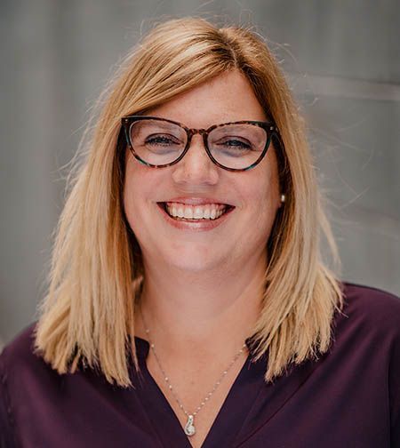 Woman with blonde hair and glasses smiles. She wears a purple shirt and a silver necklace.