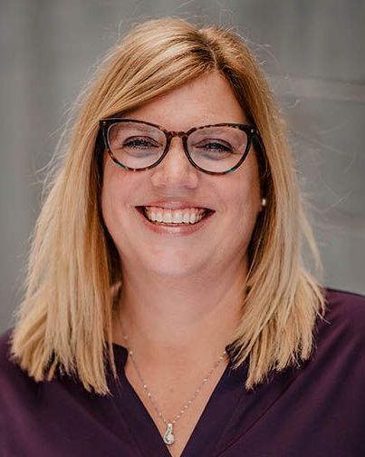 Woman with blonde hair and glasses smiles. She wears a dark purple top and a necklace.