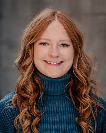 Woman with long, wavy auburn hair smiles, wearing a teal turtleneck, against a gray background.